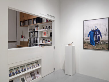 Exhibition space with a framed print on white walls and a sculpture on a plinth, with a gallery office visible beside it, featuring a bookshelf filled with books, sculptures, and framed photos.