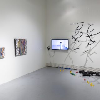 An installation photo from Access is Love and Love is Complicated. On the right, black twigs float suspended from the ceiling, a tv monitor is to the left with a scene from S/Pace visible. On the ground are more sticks and rubber toys. Turning the corner for the wall, are two touchable mosaic works, one depicting the profile of hips and the small of a back, in green, blues and orange tiles.