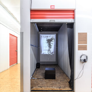 Three Windows by Aislinn Thomas installed at Holding Patterns held at a storage locker facility. The locker has a black curtain, opened to reveal a projection on a canvas of a window.