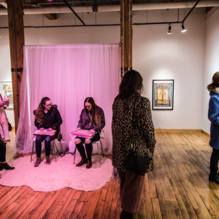 Opening reception for Outliers on Tour. A hot pink light bathes two people sitting on white chairs holding pink pillows. To their left, two people chat and to their right, someone observes an art piece from off frame.