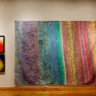 An installation shot of Outliers on Tour. A painting on canvas measuring 7x11 feet is on the right, and two smaller digital prints on the left. The work on the right is called Mountain and resembles a rainbow multicoloured band punctuated by an organic grid of squares and rectangles. To the right, the two digital prints resemble abstract watercolours.