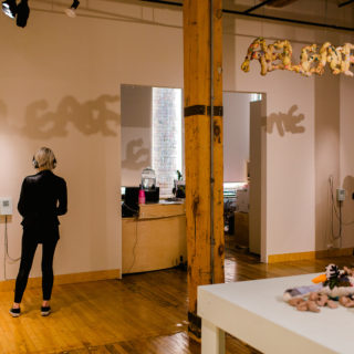 There are two people facing the back wall of Tangled Art Gallery, each dressed in black and wearing a headset. They are listening to two audio stations situated on the wall in front of them. Behind them, a soft sculpture object hangs, as if floating, between two large wooden pillars.