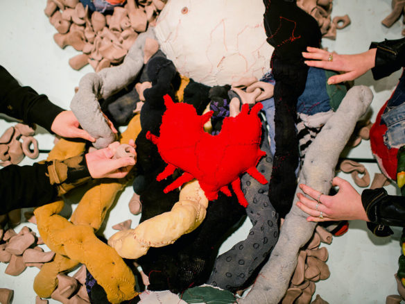 From above, a photo of two people touching a sprawling textile sculpture called Big Softie. They reach into the guts, which are soft and dimpled and made from stuffed knee socks and nylon stockings, and examine the Unidentified Remains that are scattered amongst them. At the centre lies Big Softie’s heart, a red patchwork soft sculpture with tendrils extending outward.