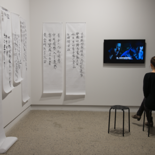 On the left, Chinese characters are roughly painted in black onto six paper scrolls of different lengths hung in a row across two walls; one scroll is long enough to pool onto the floor. A gallery visitor sits on one of two stools in front of a video monitor hung on showing two people of Asian descent looking downwards with serious expressions.