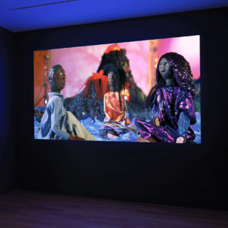 A video projected on to a wall shows three hand-made puppets sitting facing each other in a semi-circle; each puppet respectively represents one of the three Black artists who made the video.