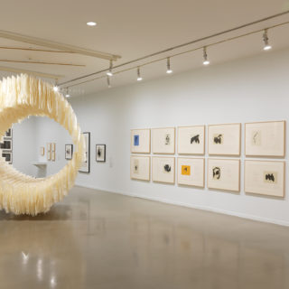 A brightly lit gallery with white walls. In the centre foreground, there is a circular sculpture made of sheets of crumpled rice paper hung from the ceiling. It appears to float in the space. On the walls are several framed works including two rows of prints on the right.