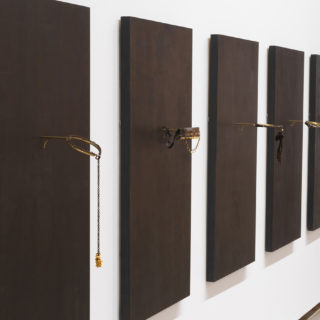 A series of five wooden panels painted black, hung on a white wall. Affixed to each panel is a circular brass armature, each of which is decorated differently, for instance with chains, corn kernels, hair, and/or leather.