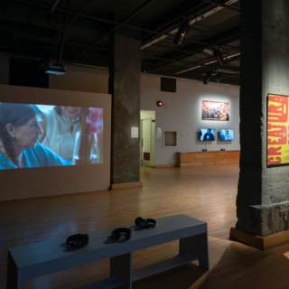 A darkly lit gallery with two concrete columns. On the left, there is a video projection showing the filmmaker Alanis Obomsawin, surrounded by two people. Her hair is in a braid and she is wearing earrings and a blue top. In front of the projection is a bench with three headphones. On the column next to the projection is poster with the title of the video “Creatura Dada” in bright red capital letters against a yellow background. On the right a black hutch holds a video monitor. In the very back, there is big banner with two video screens under it.