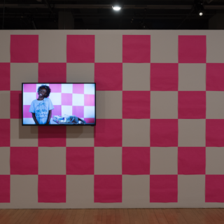 A white wall intermittently covered with large square fluorescent Post-it notes, creating a pink and white checkerboard pattern. A video monitor is hung on the left side of the wall, with headphones. The monitor shows a smiling Black pre-teen in a T-shirt that says “Super Genius,” and standing in front of a pink-and-white patterned wall like the one in the gallery.