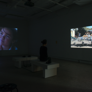 A dark gallery with two video projections on separate walls. The left projection shows an Indigenous child, and on the right, a jerry-rigged shopping cart piled with random objects. There are two benches, one of which is occupied by a visitor.