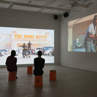 A gallery with two video projections on separate walls: one placed close to the ground while the other is higher up. There are two people in the room sitting on two of the five overturned Home Depot buckets placed around the room. The screen on the left shows five Guatemalan men all dressed in black tshirts standing or kneeling on a wooden dolly and miming shooting in various directions. They are in a Home Depot parking lot on a sunny day. In the screen on the right, a Guatemalan man in jeans and a white T-shirt brandishes his hands as if they held pointed guns.