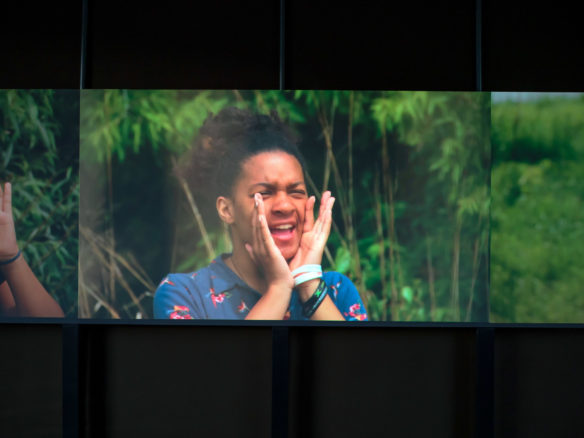 Video still of a young person of colour wearing a bright blue shirt with a floral motif, while yelling in front of a blurred backdrop of vibrant greenery.