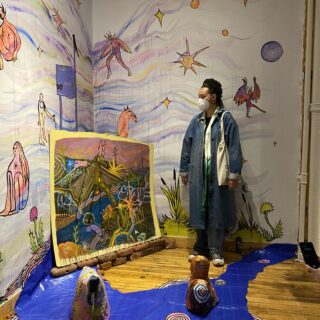 An individual wearing a long denim coat, jeans, sneakers, and a mask stands facing the artwork.
