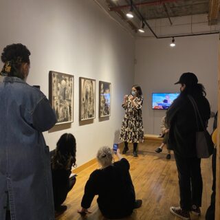 An individual with short, shoulder-length hair, wearing a black and white patterned dress, black sneakers, and a mask, stands facing artwork displayed on a white wall while a group of people gather around them, listening as they speak.