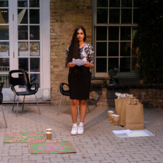 An individual with long dark hair, wearing a patterned shirt, a black skirt and white shoes speaks directly at the camera while holding a white sheet of paper.