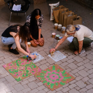 Three people crouch closely together, concentrated as they draw decorative, ornamental patterns across the bricked ground.