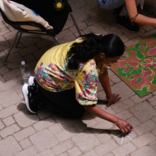An individual with their hair tied, wearing a yellow shirt with colourful flowers, black pants, and black and white shoe is crouched down, drawing a kolam pattern on the brick floor.