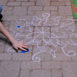 An individual sits on the brick ground, carefully colouring a kolam pattern using blue pigment.