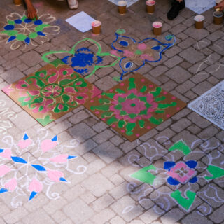 Kolam patterns are drawn across the brick floor with individuals gathered closely around drawing lines and details.