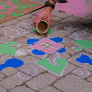 An individual crouches down, carefully filling in a kolam pattern that has been drawn on the brick floor.