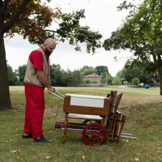 A bald, bearded individual in a red outfit and beige vest pushes a mobile table through grass with trees behind him and a house visible in the distance.