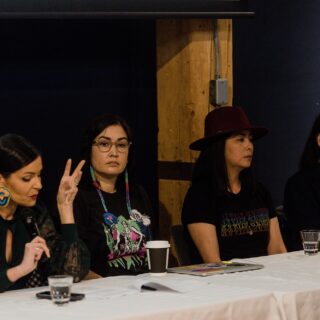 Four panelists are seated at a table. One is speaking, another is turned towards them, and the remaining two are listening attentively.