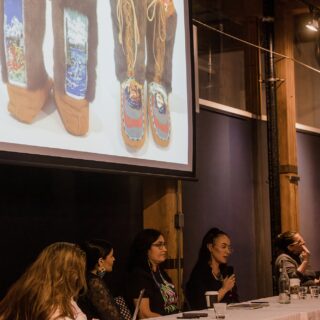 Four panelists and a moderator sit at a table, actively engaged in discussion, with a projector above them displaying an image.