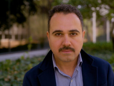 A man in a striped button-up shirt and blazer looking straight at the camera.