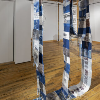 A hanging, mixed media artwork with vertically suspended fabric strips featuring blue cyanotypes, black and white images, abstract patterns, and textures.