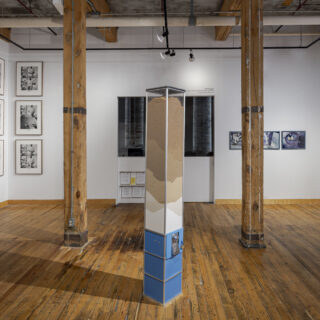 A blue candy machine containing layers of sugar stands in a gallery between wooden pillars, a series of six black and white framed photographs is to the left and a series of three framed photos is to the right.