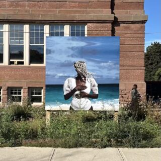 A billboard outside a brick building shows a photographic self portrait of the artist, a black woman dressed in white with a headpiece made from shells, standing on a beach with the ocean in the background.