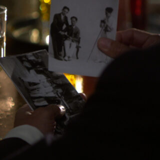 A close-up image of a man's hands, dressed in a suit, holding two black-and-white photographs. He is standing at a dimly lit bar with a half-full glass of white wine on the left side of the frame. The lighting creates a warm, reflective glow on the glass and table, adding a nostalgic and intimate atmosphere to the scene. One photograph shows two men, one seated and one standing, being photographed by a third man next to a camera on a tripod. The other image captures a group of people standing in front of a 1930s-era car.