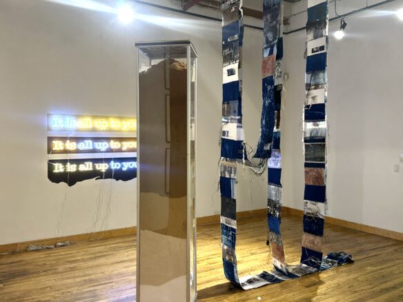 xexhibition view with three lines of neon-like text that read “It is all up to you!” on the far wall, a 6ft column made up of 5 kinds of sugar layered inside a customized candy vending machine in the foreground, and a long garland comprising photos, cyanotypes, fabric, raffia, and other images and materials stitched together and hung in three long loops from the ceiling between the column and front door