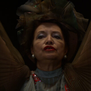 A close-up image of a singer, a woman in her late 50s, with dark hair, wearing a large decorative headpiece made of layered fabric. She has pearl earrings, bold red lipstick, and is dressed in a high-collared, pleated garment with shimmering details. The lighting casts shadows on her face, creating a dramatic and intense expression as she gazes toward her audience beyond the frame of the image. The dark background further emphasizes her face and attire.