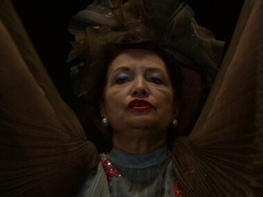 A close-up image of a singer, a woman in her late 50s, with dark hair, wearing a large decorative headpiece made of layered fabric. She has pearl earrings, bold red lipstick, and is dressed in a high-collared, pleated garment with shimmering details. The lighting casts shadows on her face, creating a dramatic and intense expression as she gazes toward her audience beyond the frame of the image. The dark background further emphasizes her face and attire.