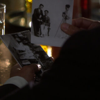 A close-up image of a man's hands, dressed in a suit, holding two black-and-white photographs. He is standing at a dimly lit bar with a half-full glass of white wine on the left side of the frame. The lighting creates a warm, reflective glow on the glass and table, adding a nostalgic and intimate atmosphere to the scene. One photograph shows two men, one seated and one standing, being photographed by a third man next to a camera on a tripod. The other image captures a group of people standing in front of a 1930s-era car.