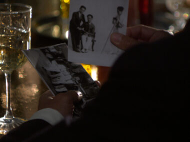 A close-up image of a man's hands, dressed in a suit, holding two black-and-white photographs. He is standing at a dimly lit bar with a half-full glass of white wine on the left side of the frame. The lighting creates a warm, reflective glow on the glass and table, adding a nostalgic and intimate atmosphere to the scene. One photograph shows two men, one seated and one standing, being photographed by a third man next to a camera on a tripod. The other image captures a group of people standing in front of a 1930s-era car.