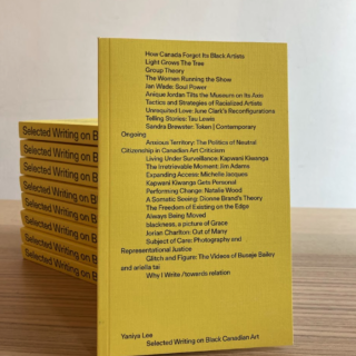 paperback book with yellow cover featuring list of essay titles in black type, leaned up against a stack of the same book