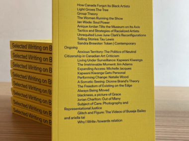 paperback book with yellow cover featuring list of essay titles in black type, leaned up against a stack of the same book