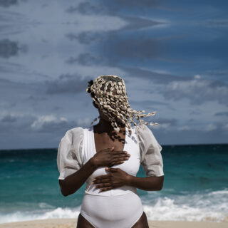 A woman with dark brown skin is shown from the hips up standing on a beach in front of rolling ocean waves. She wears a white bodysuit with bloused sleeves, her hands placed over heart and abdomen, and her face is obscured by a covering beaded in cowrie shells.