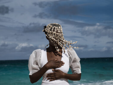 A woman with dark brown skin is shown from the hips up standing on a beach in front of rolling ocean waves. She wears a white bodysuit with bloused sleeves, her hands placed over heart and abdomen, and her face is obscured by a covering beaded in cowrie shells.