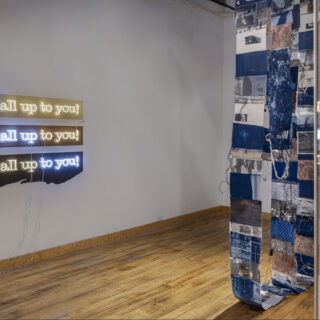 [Installation view of an art exhibition featuring a LED neon artwork with the words “It is all up to you” in yellow and blue, and a hanging mixed-media piece with blue, black, and white photographic prints stitched together