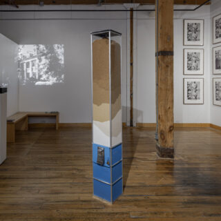 A blue candy machine containing layers of sugar stands in a gallery with a black and white projected image to the left and a series of six black and white framed photographs to the right