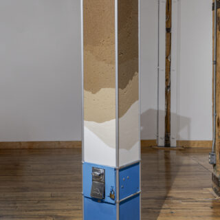 A blue candy machine containing layers of sugar in various shades of brown and white stands on a wooden floor in front of a white wall in a gallery.