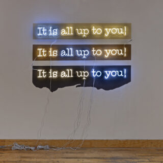 Installation view of an artwork with the words “It is all up to you” repeated three times in blue and yellow LED neon, on three brown painted backgrounds of varying shades.