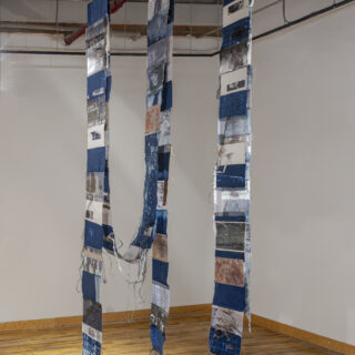 A hanging, mixed media artwork with vertically suspended fabric strips featuring blue cyanotypes, black and white images, abstract patterns, and textures.