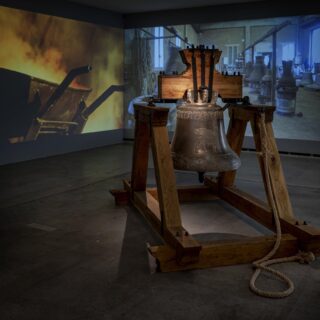 A large bell in front of a two-channel video projection depicting a foundry