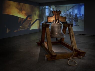 A large bell in front of a two-channel video projection depicting a foundry