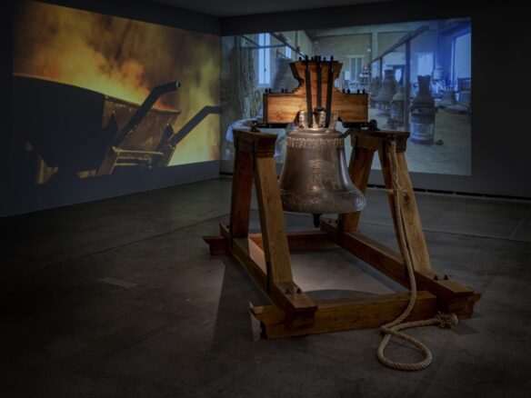 A large bell in front of a two-channel video projection depicting a foundry