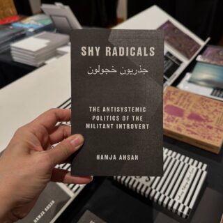 A light brown-skinned hand holds a black, softcover book titled "Shy Radicals".